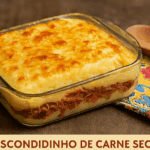 Escondidinho de carne seca em uma travessa de vidro, com recheio de carne desfiada, purê de aipim e uma crosta de queijo gratinado por cima.