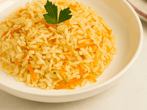 Arroz com cenoura ralada soltinho, servido em formato de montinho em um prato fundo branco e finalizado com uma folha de salsinha fresca por cima.