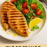 Dois filés de frango grelhado com marcas de grelha escuras, servidos em um prato branco ao lado de uma salada fresca de alface, tomate cereja e cenoura ralada, com uma fatia de limão.