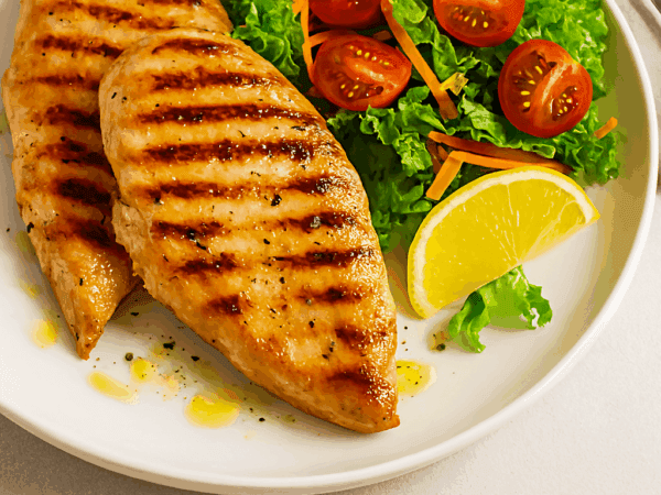 Dois filés de frango grelhado com marcas de grelha escuras, servidos em um prato branco ao lado de uma salada fresca de alface, tomate cereja e cenoura ralada, com uma fatia de limão.
