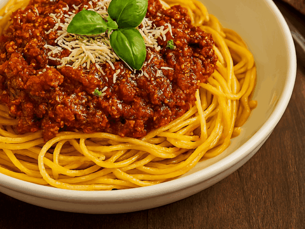 Macarrão à bolonhesa servido em um prato fundo branco, com o espaguete coberto por um molho de carne moída espesso, queijo parmesão ralado e folhas frescas de manjericão.