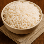 Uma tigela redonda de cor clara cheia de arroz branco soltinho, servida sobre um guardanapo de linho em uma mesa de madeira escura.