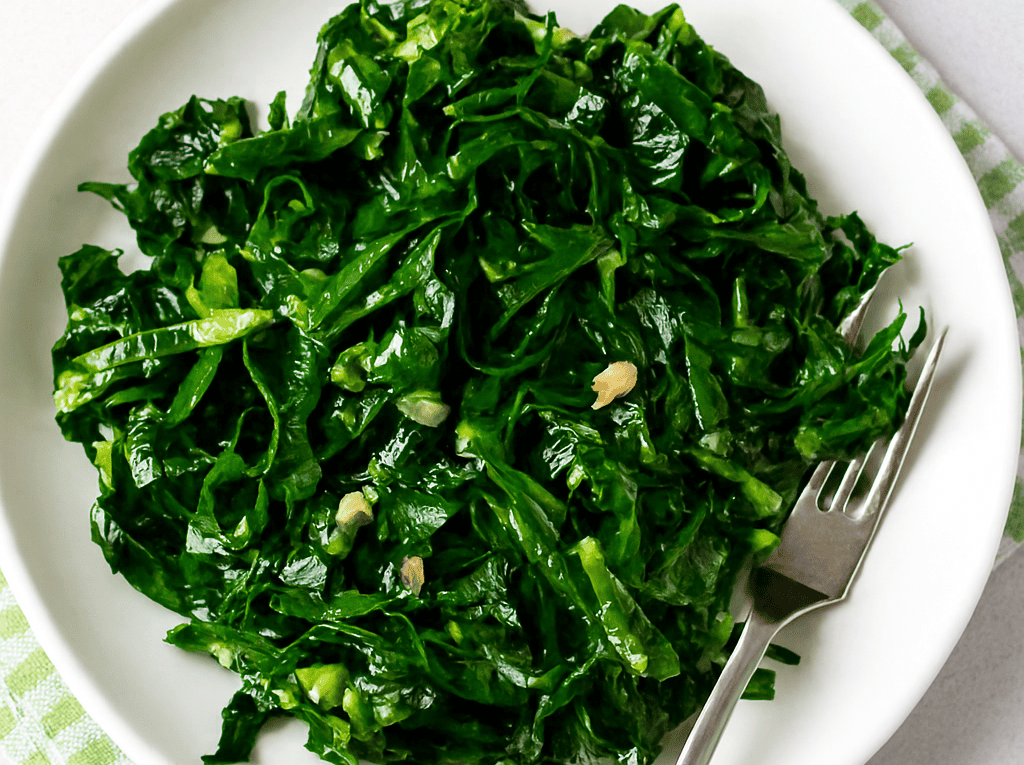 Uma porção de couve refogada com alho, bem verdinha e brilhante, servida em um prato branco redondo com dentes de alho picados e uma garfo ao lado.