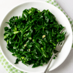 Uma porção de couve refogada com alho, bem verdinha e brilhante, servida em um prato branco redondo com dentes de alho picados e uma garfo ao lado.