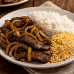 Iscas de fígado aceboladas com bastante cebola dourada, servidas em um prato branco ao lado de arroz branco soltinho e farofa amarela.