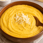Polenta cremosa servida em uma tigela marrom rústica, finalizada com um montinho de queijo ralado e pimenta-do-reino, com uma colher de madeira mergulhada no prato.