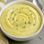 Purê de batata bem cremoso servido em uma tigela branca redonda, com a superfície decorada em espiral e salpicada com cebolinha picada e pimenta.