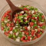Vinagrete clássico para churrasco em uma tigela de vidro, com tomate, cebola e pimentão picados em cubinhos, sendo misturado com uma colher de madeira.