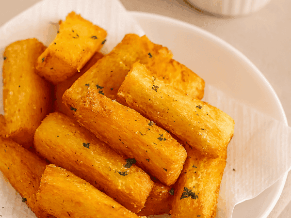 Porção de aipim frito na manteiga de garrafa, bem dourado e crocante, servido em um prato branco sobre um guardanapo de papel, com um potinho de molho ao fundo.
