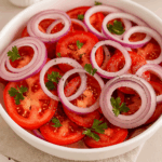 Salada de tomate em rodelas com anéis de cebola roxa servida em uma tigela branca, decorada com folhas de salsa fresca e temperada com pimenta-do-reino.