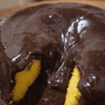 Bolo de cenoura vulcão redondo, coberto por uma camada espessa e brilhante de calda de chocolate que escorre pelas laterais e pelo centro, lembrando lava. Uma fatia foi retirada, revelando a massa amarela e úmida. O bolo está sobre um prato de vidro, com fundo desfocado em tons quentes que sugerem ambiente de cozinha.