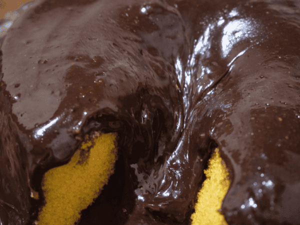 Bolo de cenoura vulcão redondo, coberto por uma camada espessa e brilhante de calda de chocolate que escorre pelas laterais e pelo centro, lembrando lava. Uma fatia foi retirada, revelando a massa amarela e úmida. O bolo está sobre um prato de vidro, com fundo desfocado em tons quentes que sugerem ambiente de cozinha.