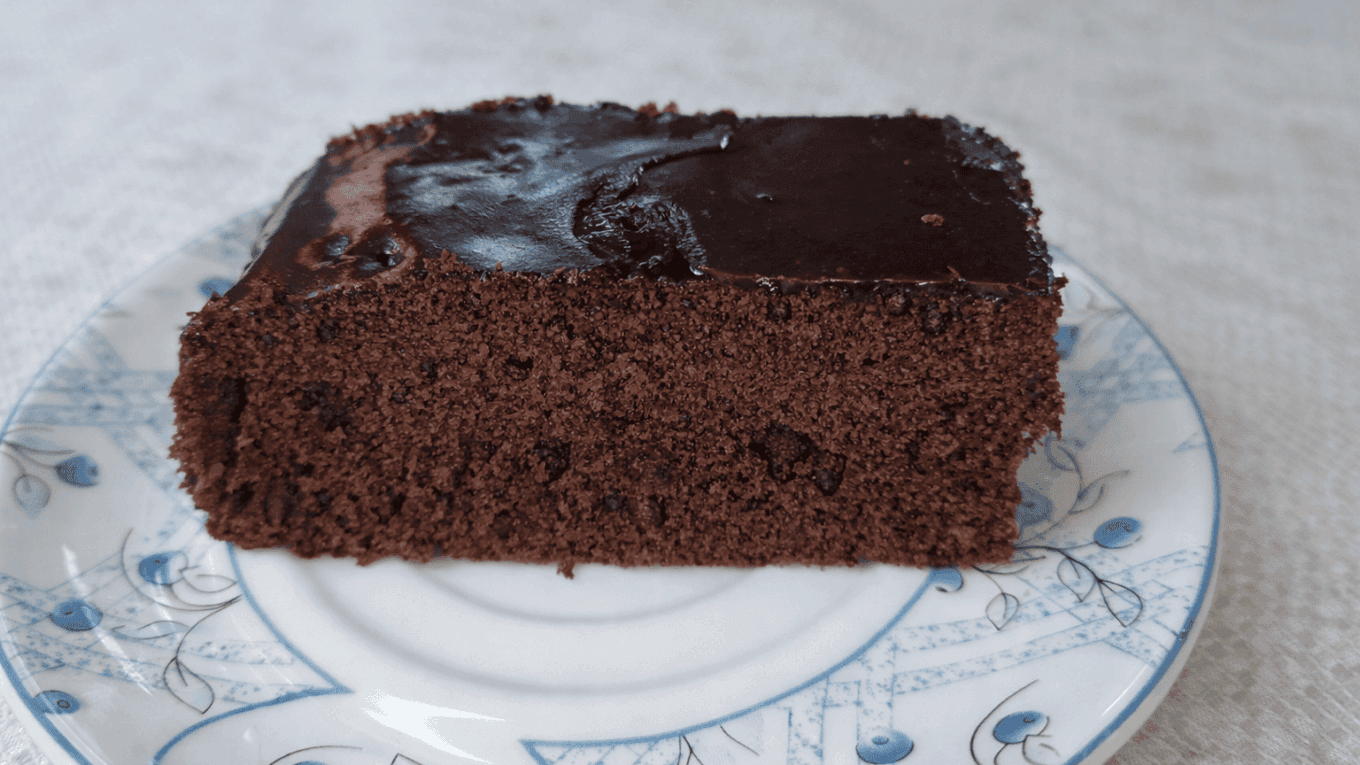 Fatia de bolo de chocolate sobre prato de porcelana branca com detalhes azuis, mostrando textura úmida e cobertura brilhante de chocolate sob luz natural.
