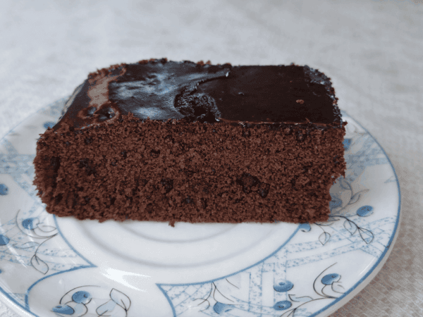 Fatia de bolo de chocolate sobre prato de porcelana branca com detalhes azuis, mostrando textura úmida e cobertura brilhante de chocolate sob luz natural.