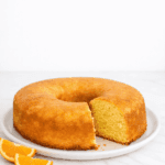Fatia fofinha de bolo de laranja recém-cortada, assado em forma redonda com furo central, destacando a textura úmida da massa e a casquinha brilhante da calda açucarada.