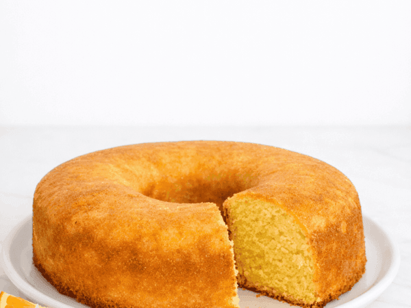 Fatia fofinha de bolo de laranja recém-cortada, assado em forma redonda com furo central, destacando a textura úmida da massa e a casquinha brilhante da calda açucarada.