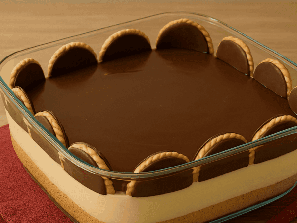 Travessa de vidro exibindo as camadas da Torta Holandesa: base crocante de biscoito, creme branco aveludado, cobertura brilhante de ganache de chocolate e laterais decoradas com biscoitos redondos Calipso.